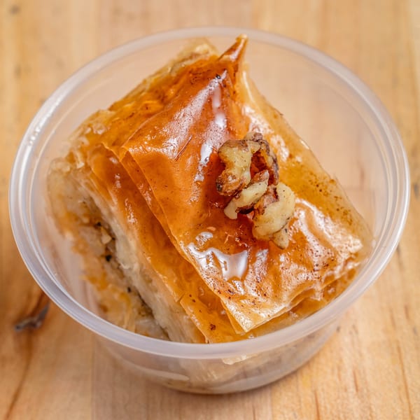 Golden brown baklava with walnuts and syrup in a clear plastic container on a wooden surface.